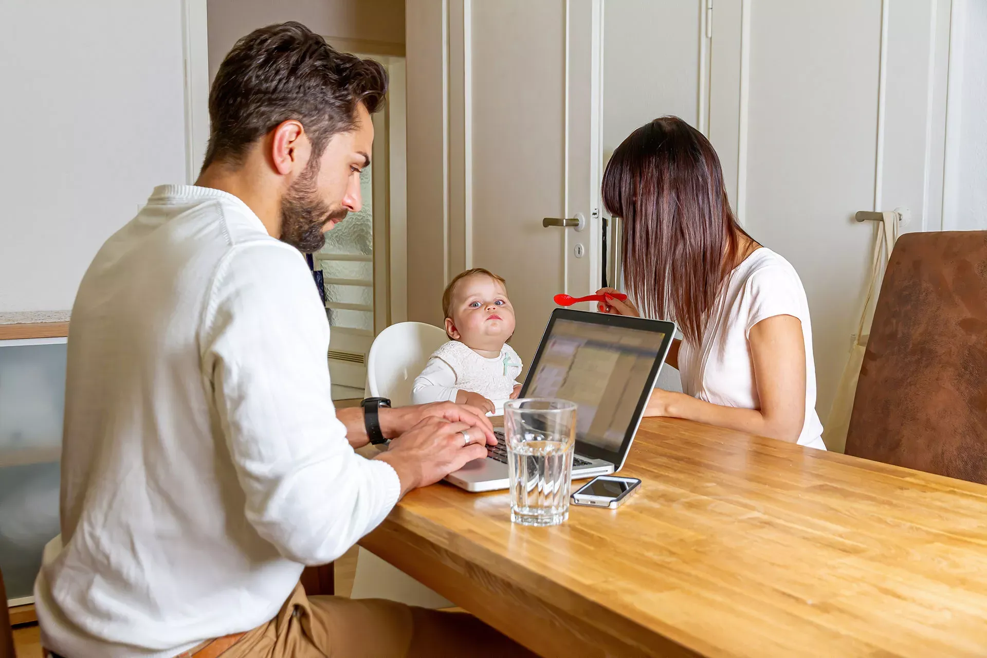 Junges Paar zuhause mit Baby am Esstisch, Mann arbeitet am Laptop im Homeoffice.. Junges Paar zuhause mit Baby am Esstisch, Mann arbeitet am Laptop im Homeoffice..