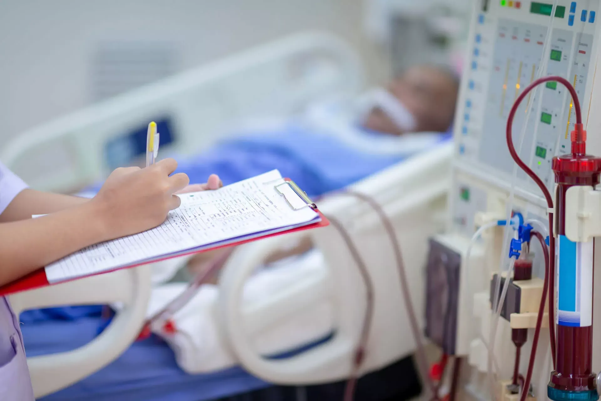 Krankenschwester überträgt Werte von einer Dialysemaschine in eine Tabelle auf einem Klemmbrett, im Hintergrund liegt der Dialysepatient im Bett. Krankenschwester überträgt Werte von einer Dialysemaschine in eine Tabelle auf einem Klemmbrett, im Hintergrund liegt der Dialysepatient im Bett.