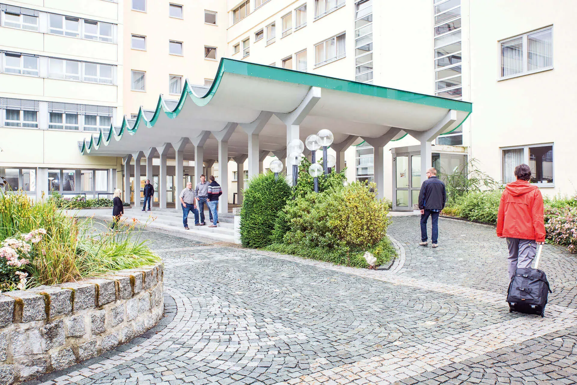 Psychosomatische Rehaklinik: Höhenklinik Bischofsgrün, Eingangsbereich mit überdachtem Portal und Menschen mit Gepäck. Psychosomatische Rehaklinik: Höhenklinik Bischofsgrün, Eingangsbereich mit überdachtem Portal und Menschen mit Gepäck.