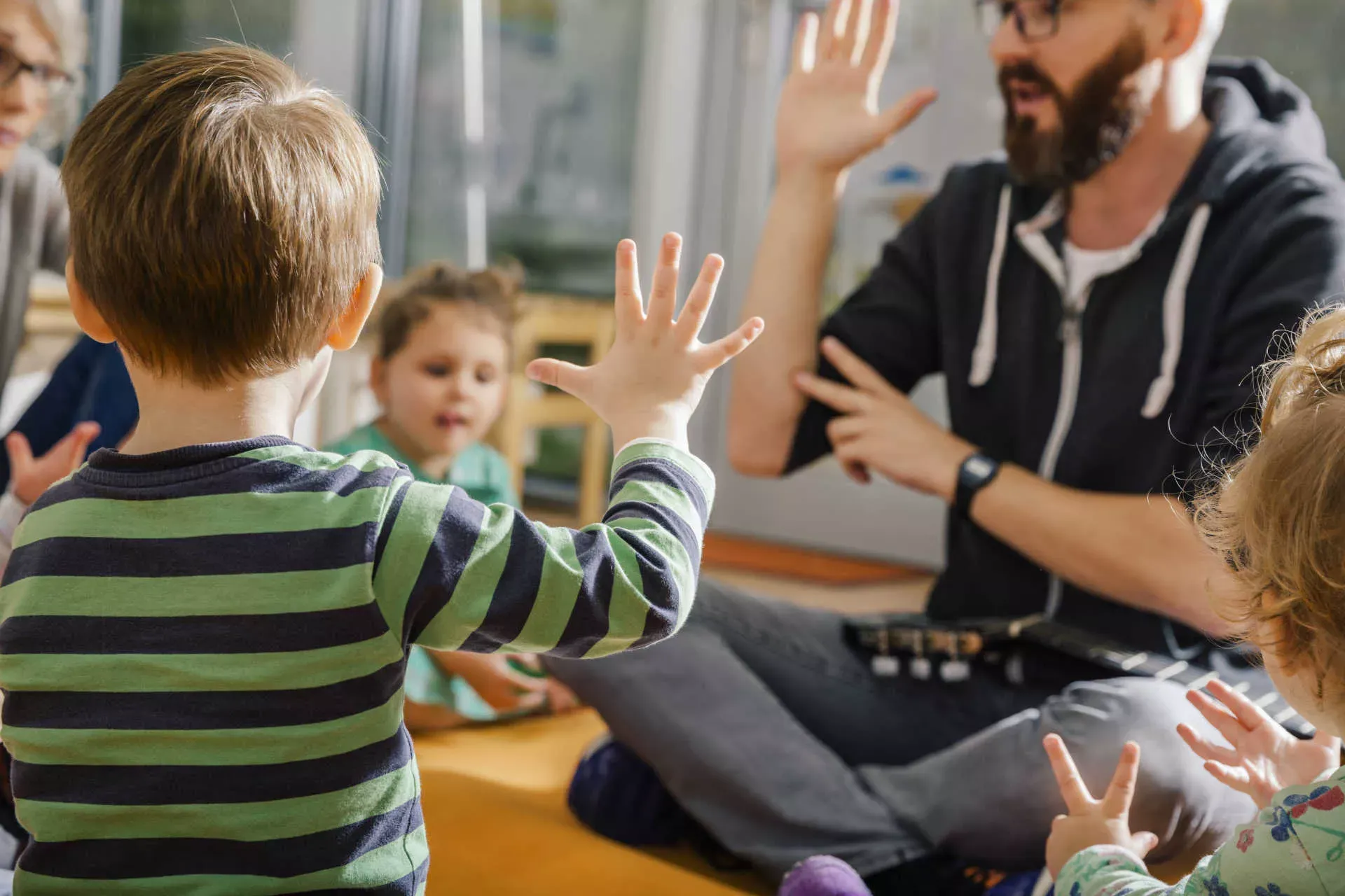 Erzieher spielt im Kreis mit Kindern in einer Kita. Bild: IMAGO / Westend61 / Mareen Fischinger