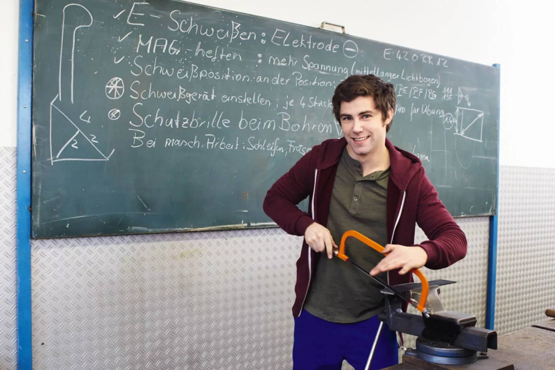 Beschreibung: Männlicher Azubi steht in der Berufsschule vor einer Tafel und sägt an einem Metallwerkstück. Männlicher Azubi steht in der Berufsschule vor einer Tafel und sägt an einem Metallwerkstück.