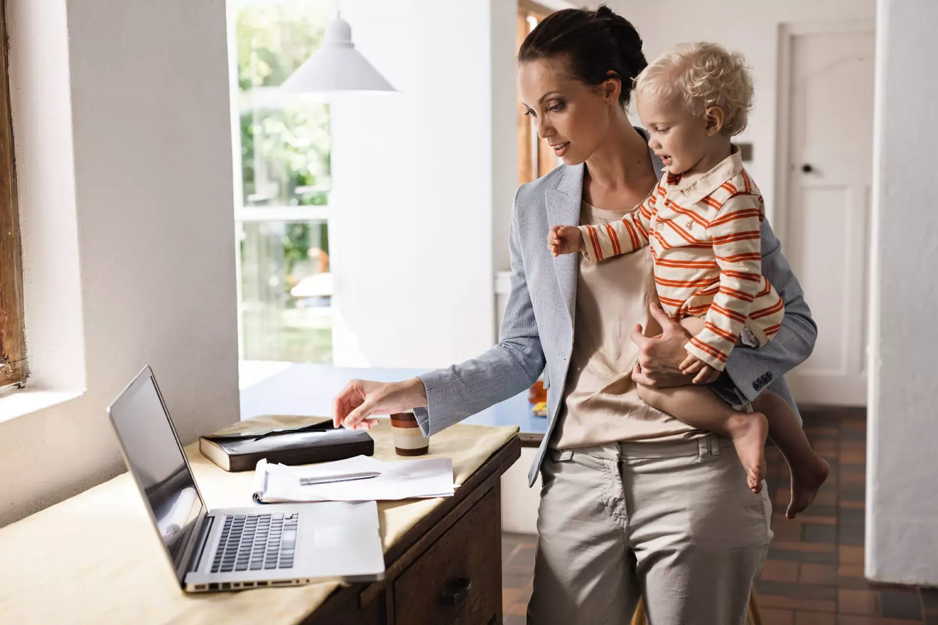 Mutter steht in Businesskleidung mit ihrem Kleinkind auf dem Arm vor einem Laptop. Mutter steht in Businesskleidung mit ihrem Kleinkind auf dem Arm vor einem Laptop.