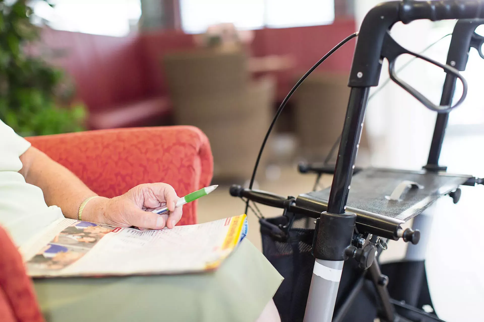 Seniorina sitzt neben ihrem Rollator auf einem Sessel mit einem Stift in der Hand und einem Kreuzworträtsel auf dem Schoß.