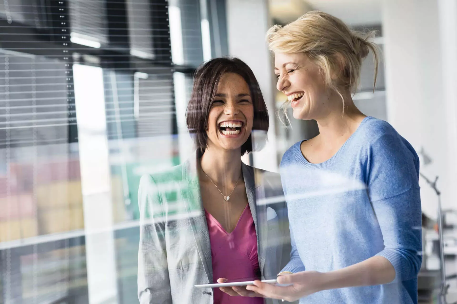 Zwei Kolleginnen stehen im Büro und lachen, eine hält ein Tablet in den Händen. Zwei Kolleginnen stehen im Büro und lachen, eine hält ein Tablet in den Händen.