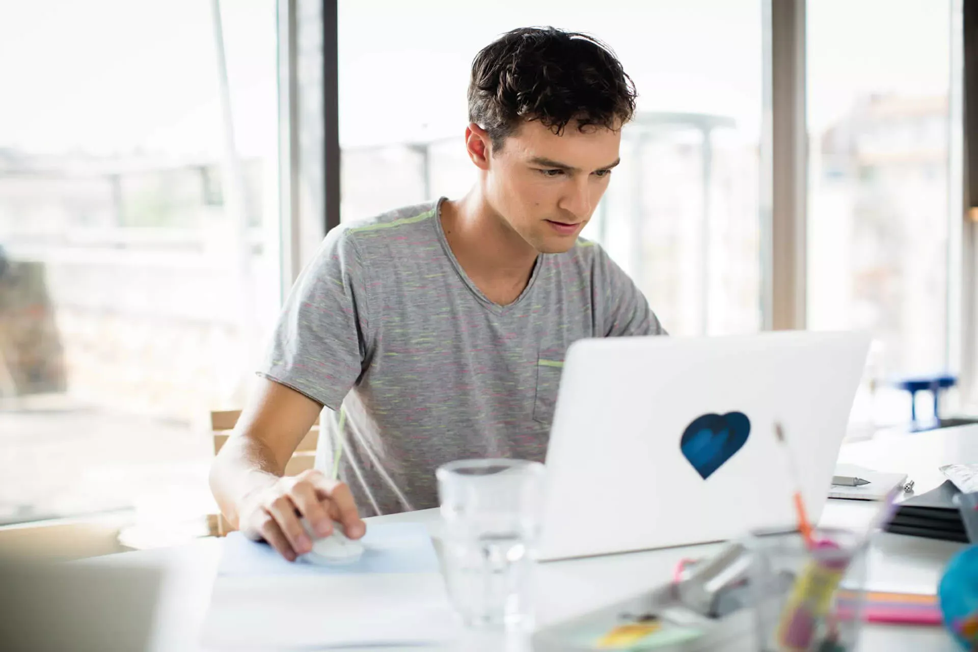 Junger Mann sitzt zuhause am Notebook mit einem Glas Wasser auf dem Tisch.
